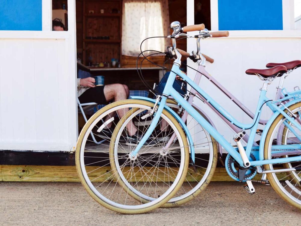 Two bikes one pink and one blue are parked outside an occupied beach hut whilst person chills inside with a cup of tea