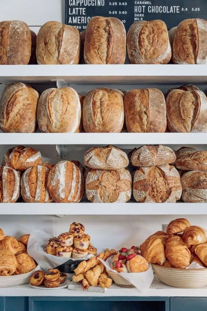 Underneath a menu board showing a selection of hot drinks, are four white shelves presented with large crusty loaves and the lowest shelf stacked with small sweet and savoury pastries